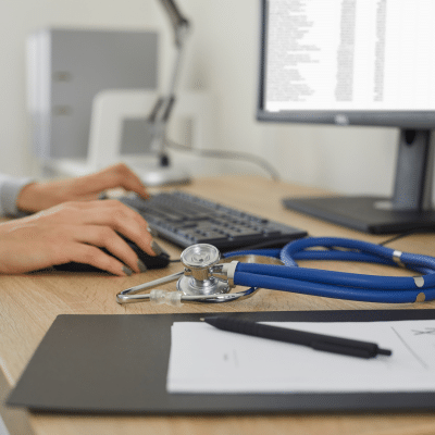 Person working on a desktop computer with clipboard and stethoscope in the foreground, representing electronic health records